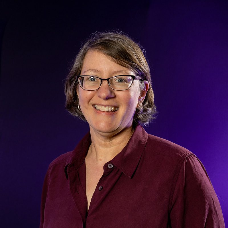 Susan Bruxvoort Lipscomb wearing red button up shirt standing in front of purple backdrop.
