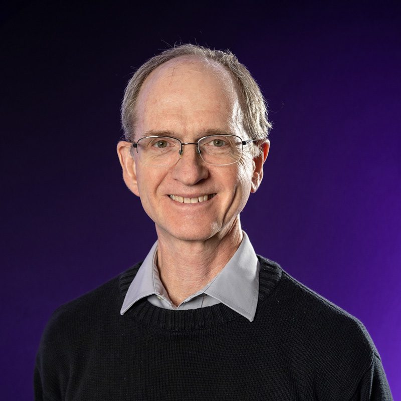 Marcus Dean wearing black sweater standing in front of purple backdrop.