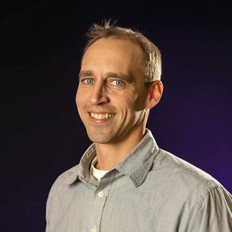 Eli Knapp wearing a grey button up shirt standing in front of purple backdrop.