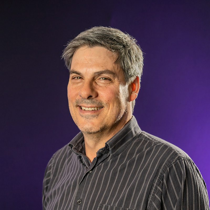 Paul Martino wearing striped button up shirt standing in front of purple backdrop.