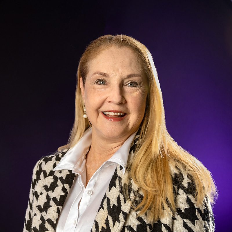 Sara Massey wearing a white button up shirt and patterned blazer standing in front for purple backdrop.