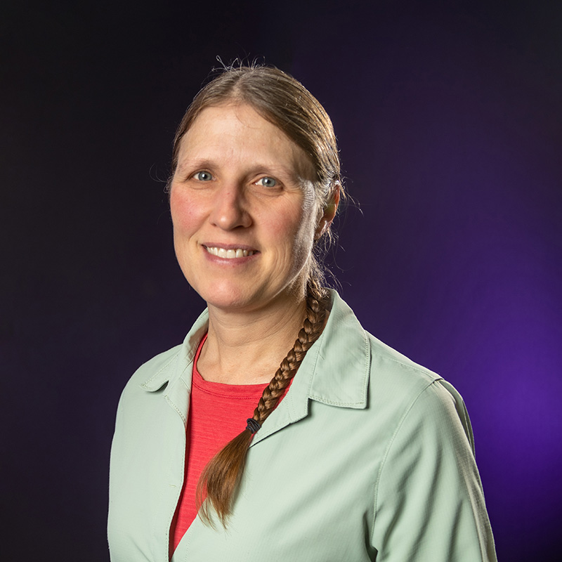 Karen Toracca wearing read shirt and green blazer standing in front of purple backdrop.