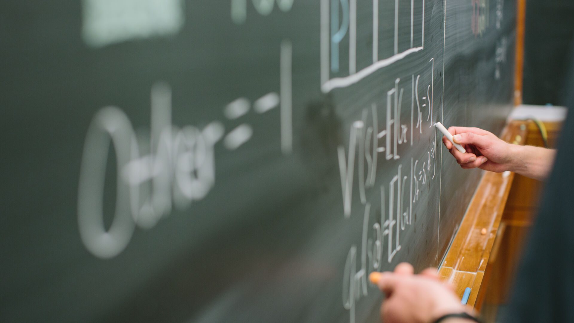 hands holding chalk and writing an equation on chalkboard