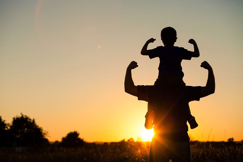 boy sitting on man's back at sunrise with arms up