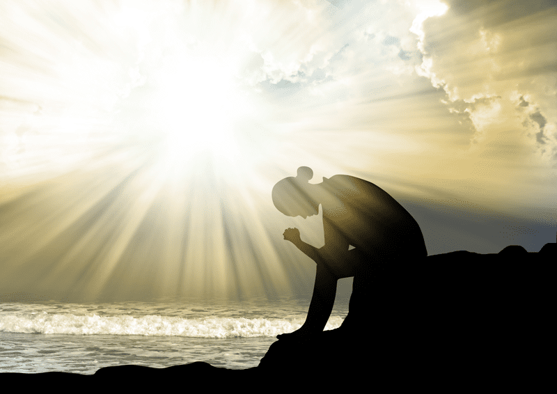 Silhouette of woman bowing head in prayer as sun shines over coast behind her