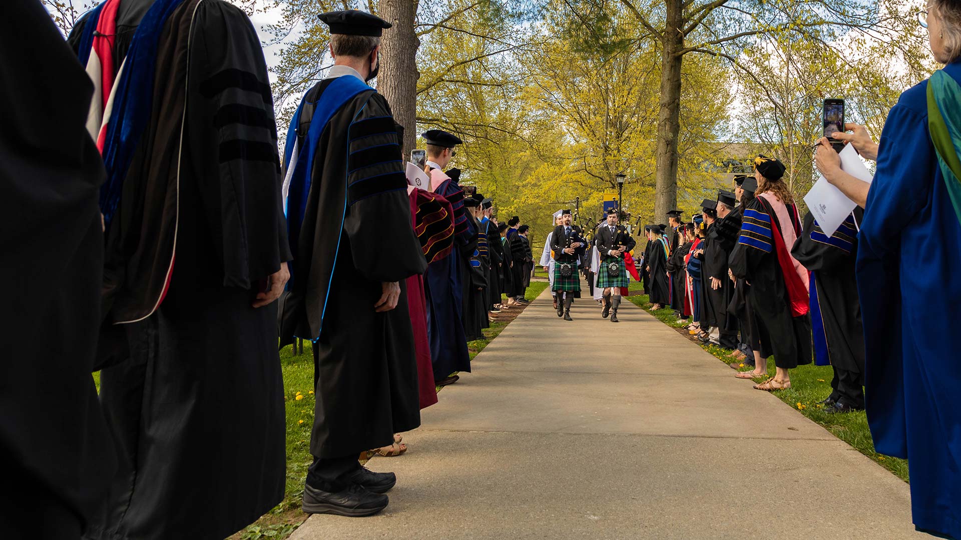 Bagpipers lead graduates down sidewalk between Houghton faculty at commencement