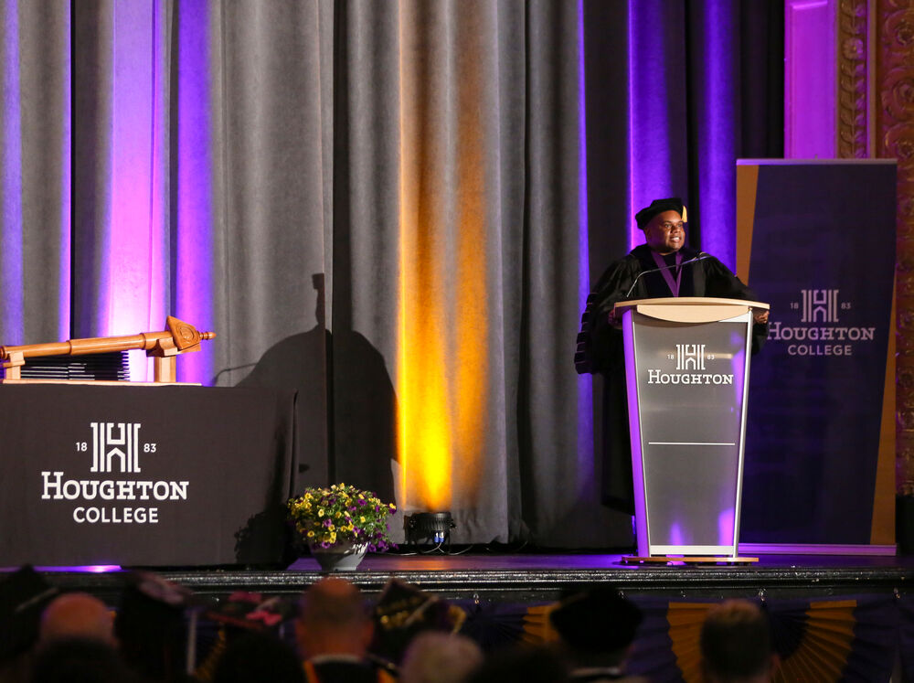 President Lewis delivering commencement address on the stage of Forbes Theater with purple and gold lights in the background