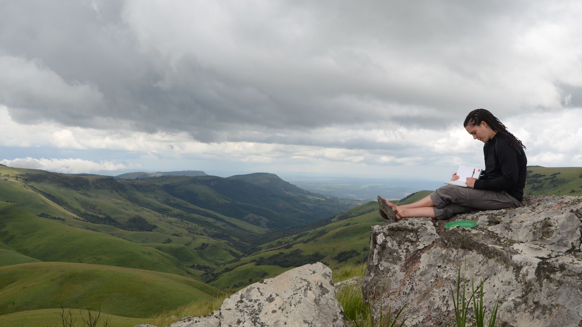Student sitting on top of cliff overlooking a scenic valley while writing in their notebook.