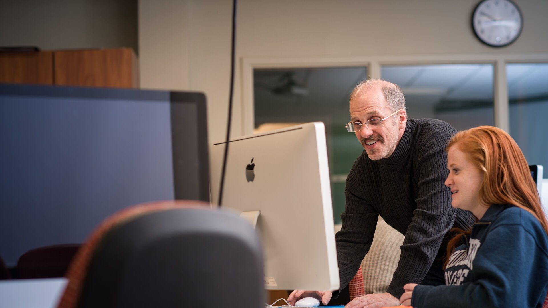 Houghton student working at Mac in Mac computer lab with professor.