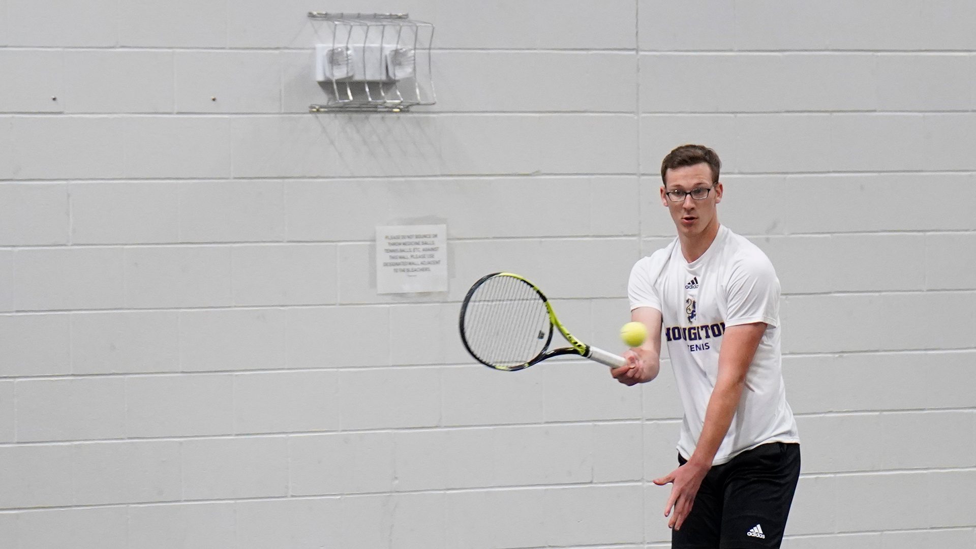 Houghton student Isaac Little hitting tennis ball on indoor court.