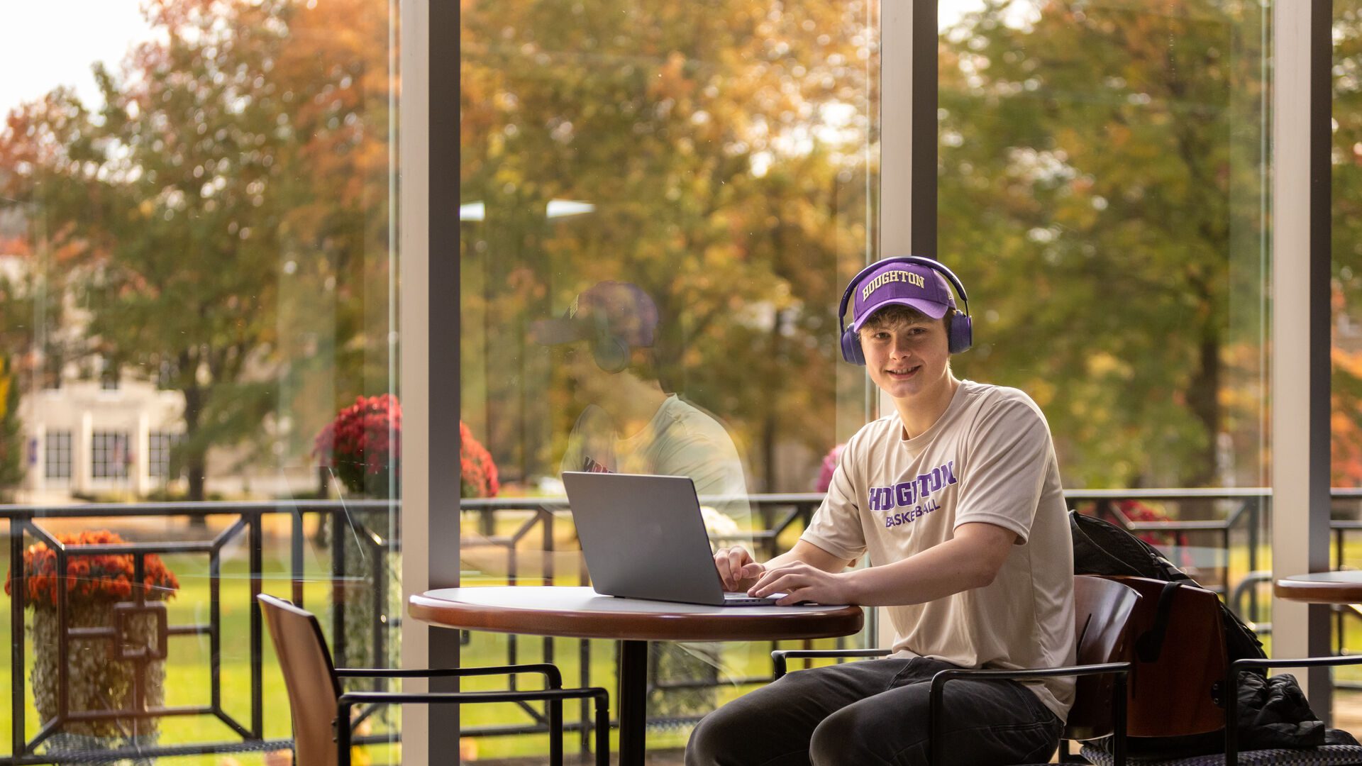 Houghton student sitting at table with laptop, wearing Houghton hat and tshirt.