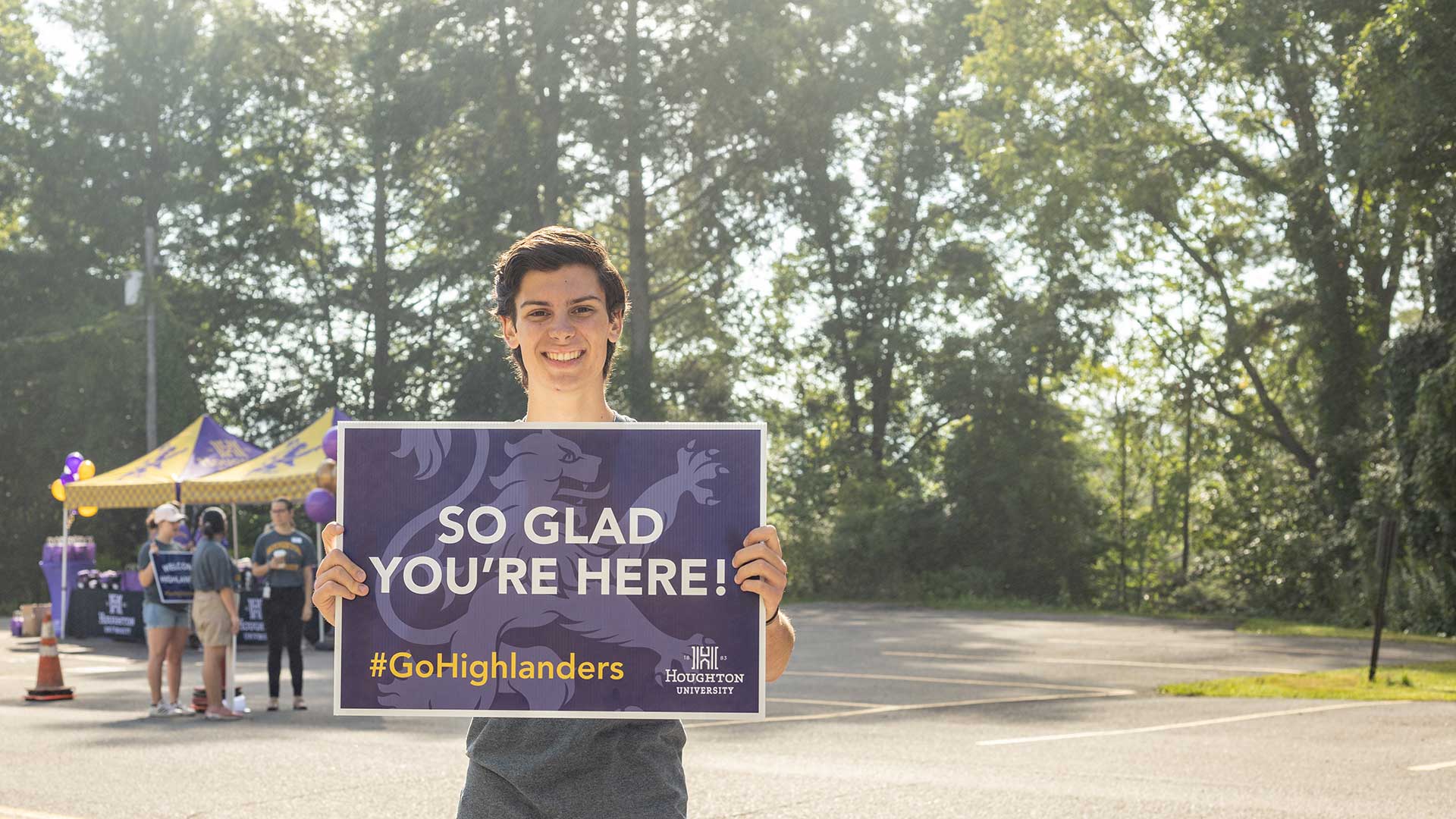 Houghton student standing with purple sign that reads, So Glad You're Here.