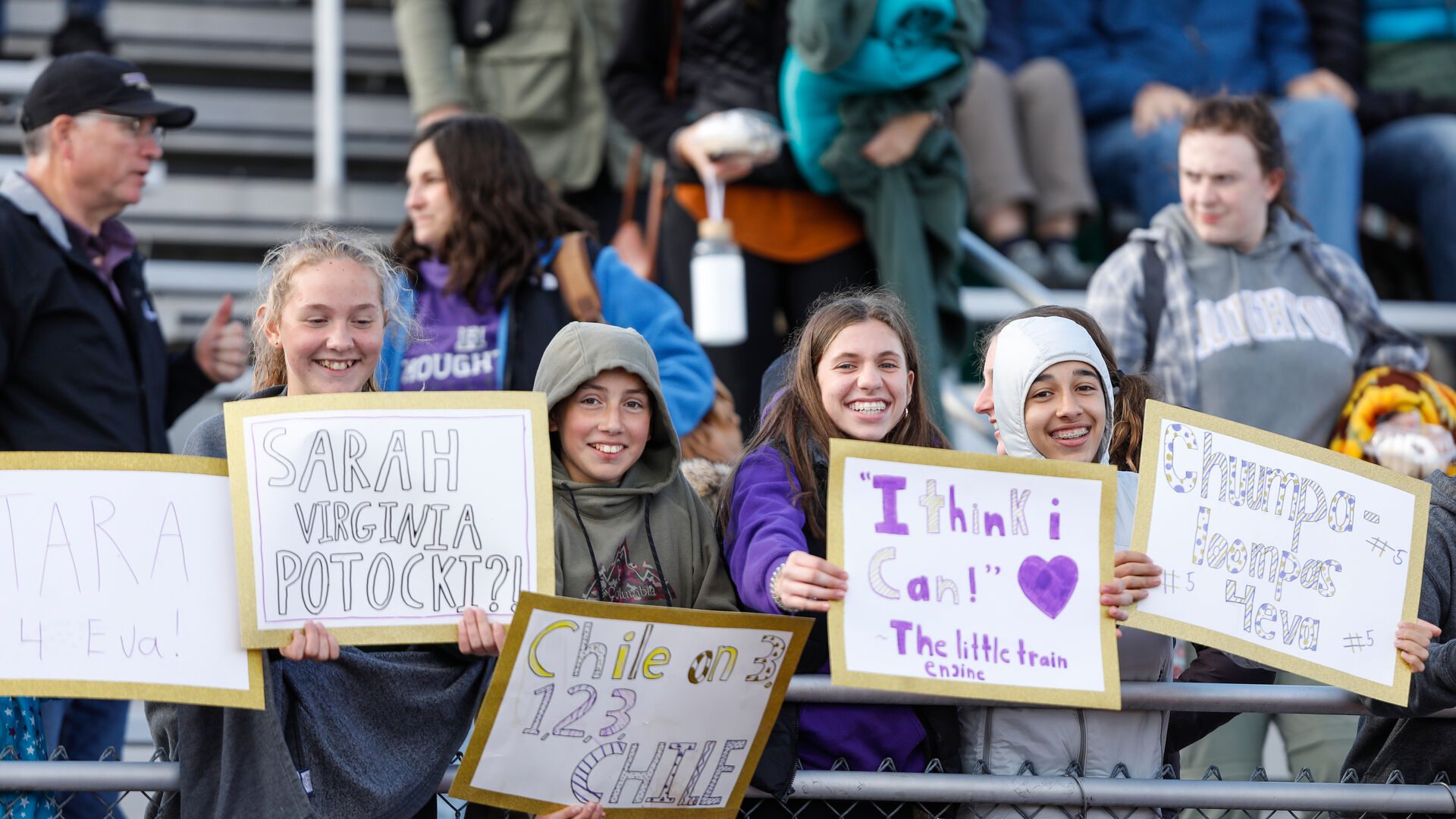 Four young ladies standing on bleachers with signs during Houghton Homecoming sporting event.