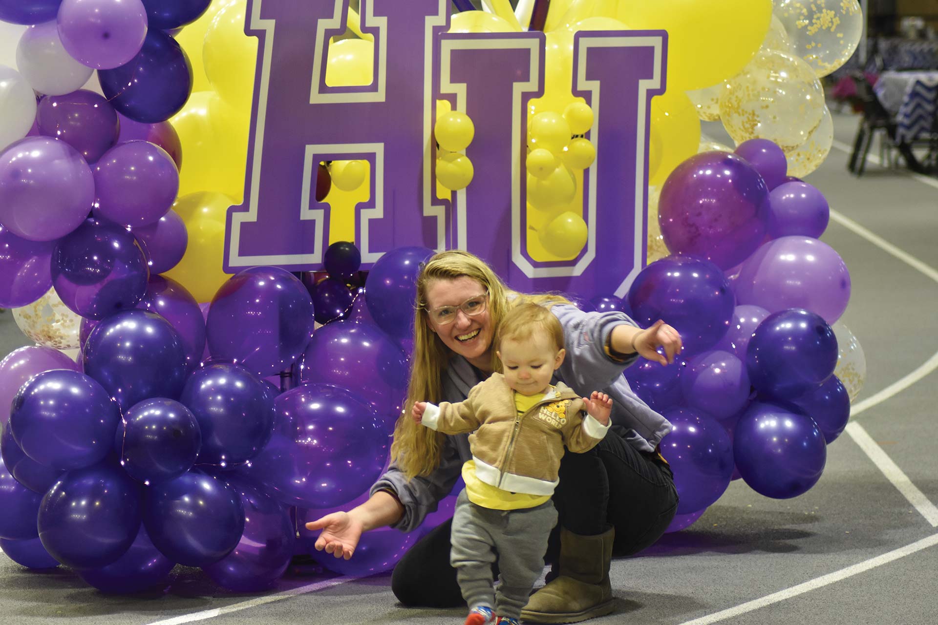 Mom with child standing in from of purple and gold balloons and HU letters at homecoming.