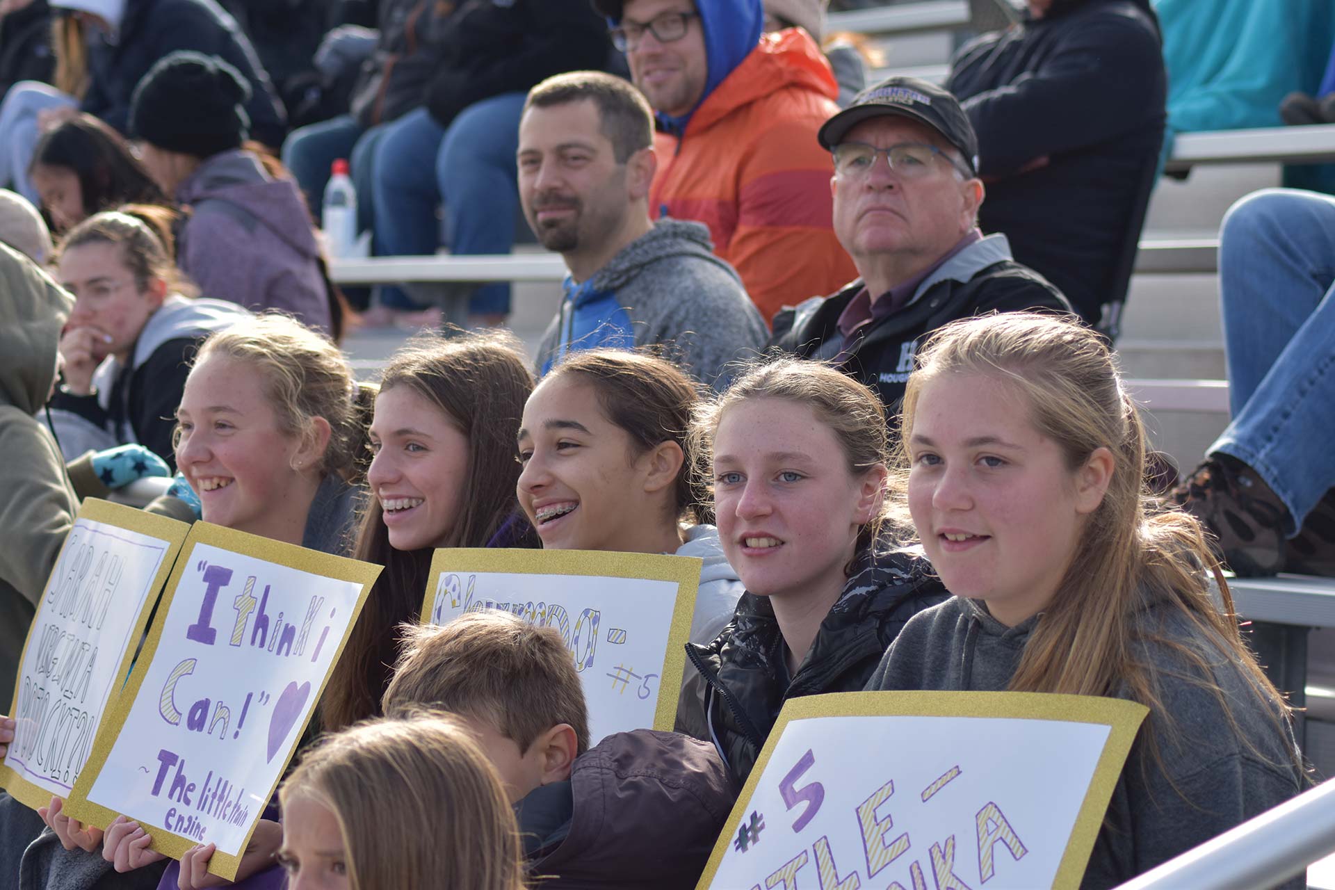 A group of young girls sitting together with signs at a sporting event during homecoming weekend at Houghton.
