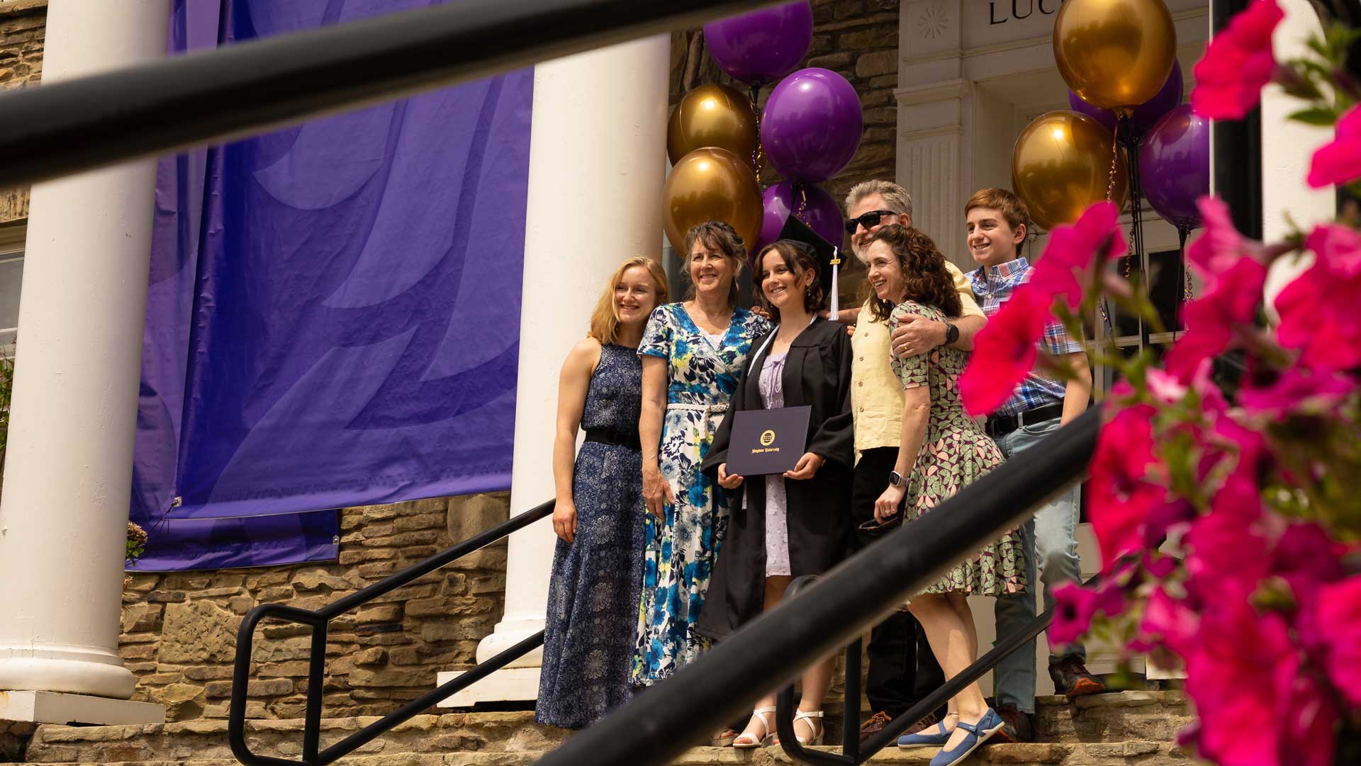 Houghton graduate and family take a photo on the steps of Luckey Memorial with balloons and flowers