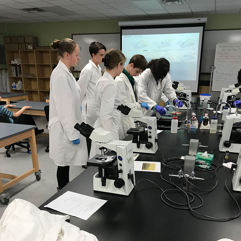 students in lab coats gathered around a microscope