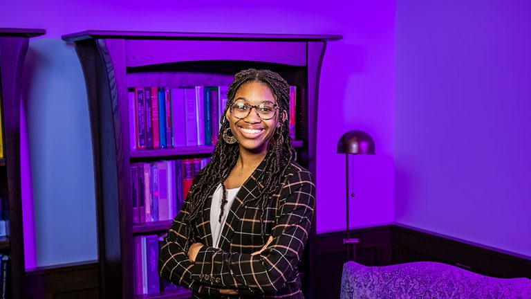 Houghton student Alexia Patterson standing in front of bookshelf wearing plaid blazer and glasses.