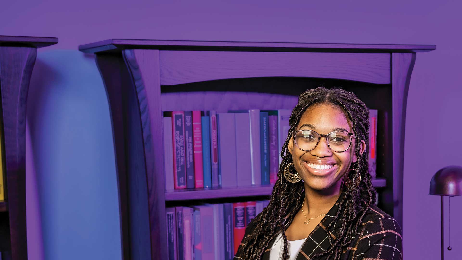 Houghton student Alexia Patterson standing in front of bookshelf wearing plaid blazer and glasses.