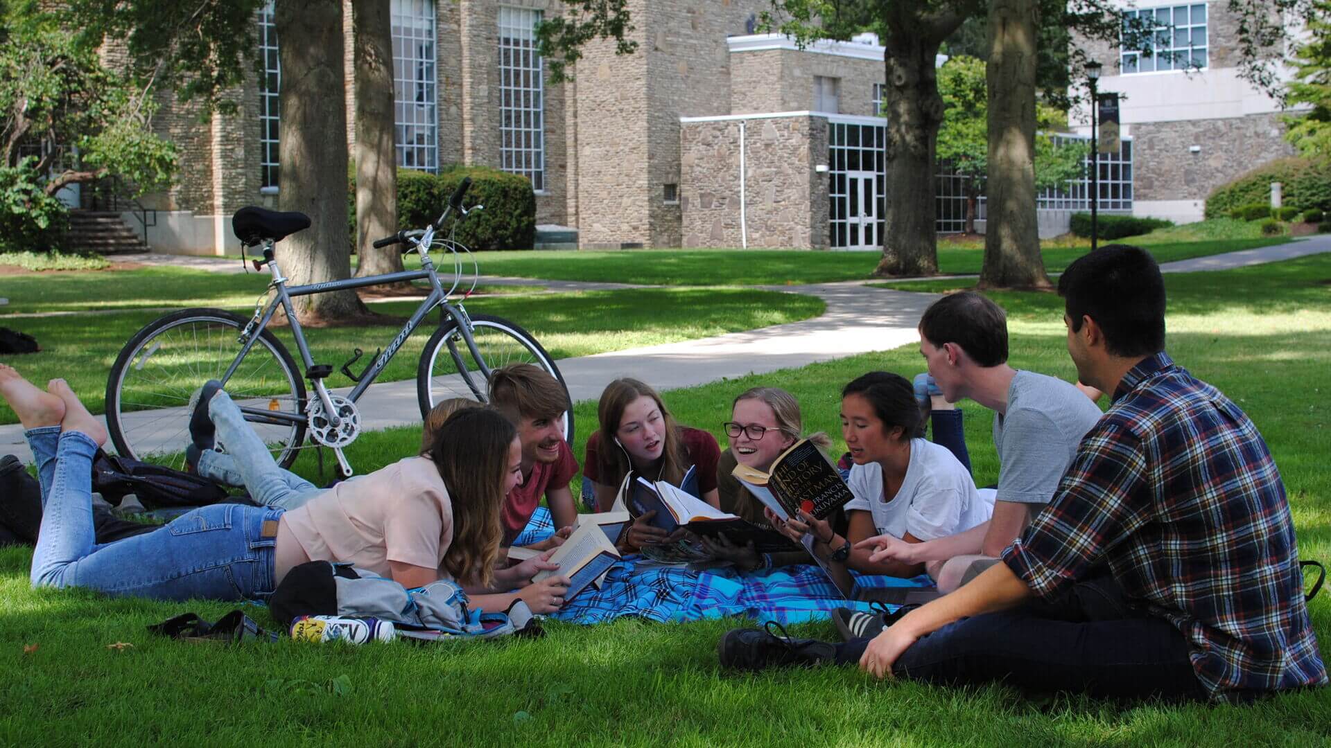 A group of Houghton students sitting on the ground on the campus lawn discussing a book.