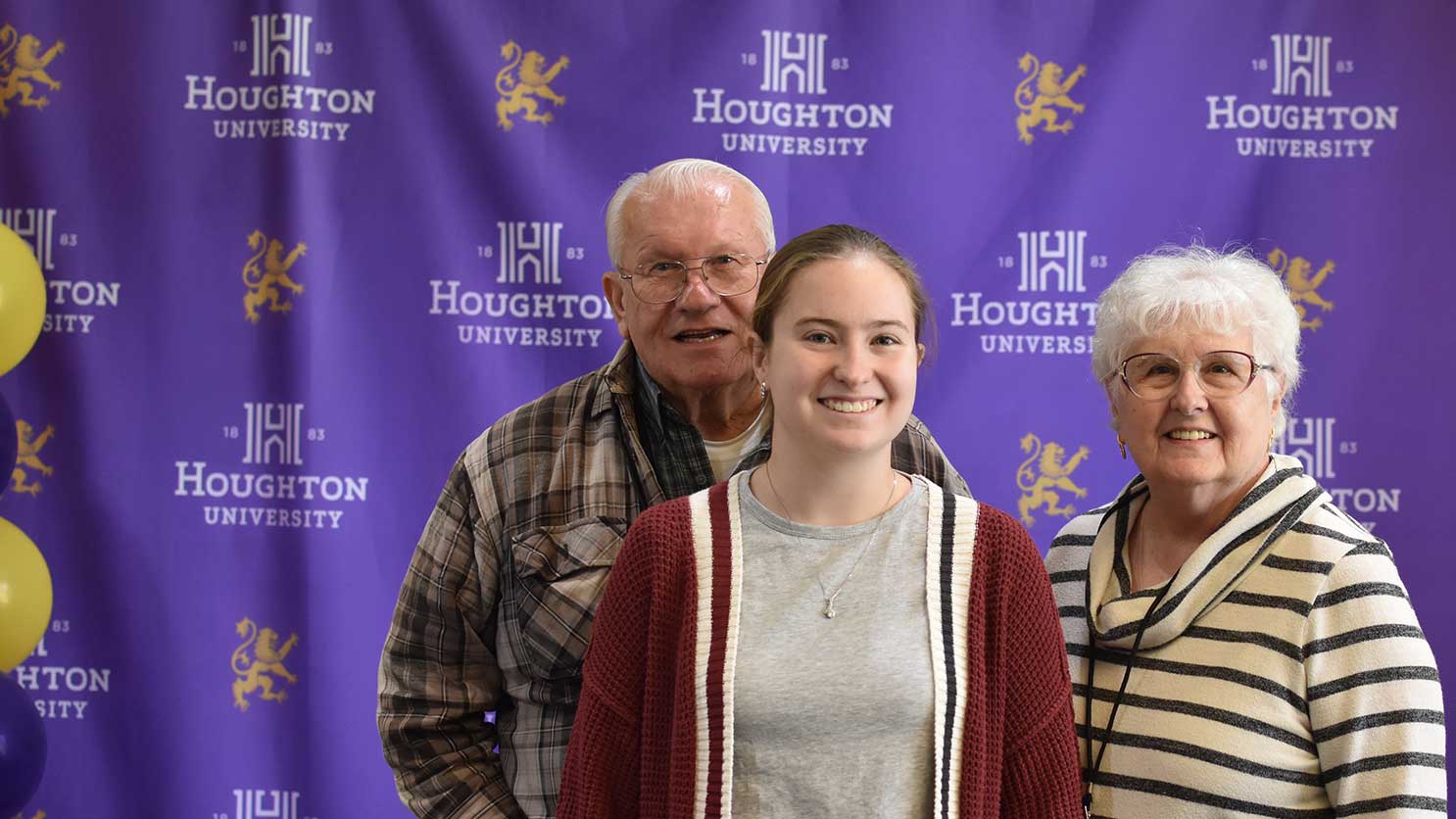 Houghton student standing with her grandparents in front of a Houghton step and repeat banner.