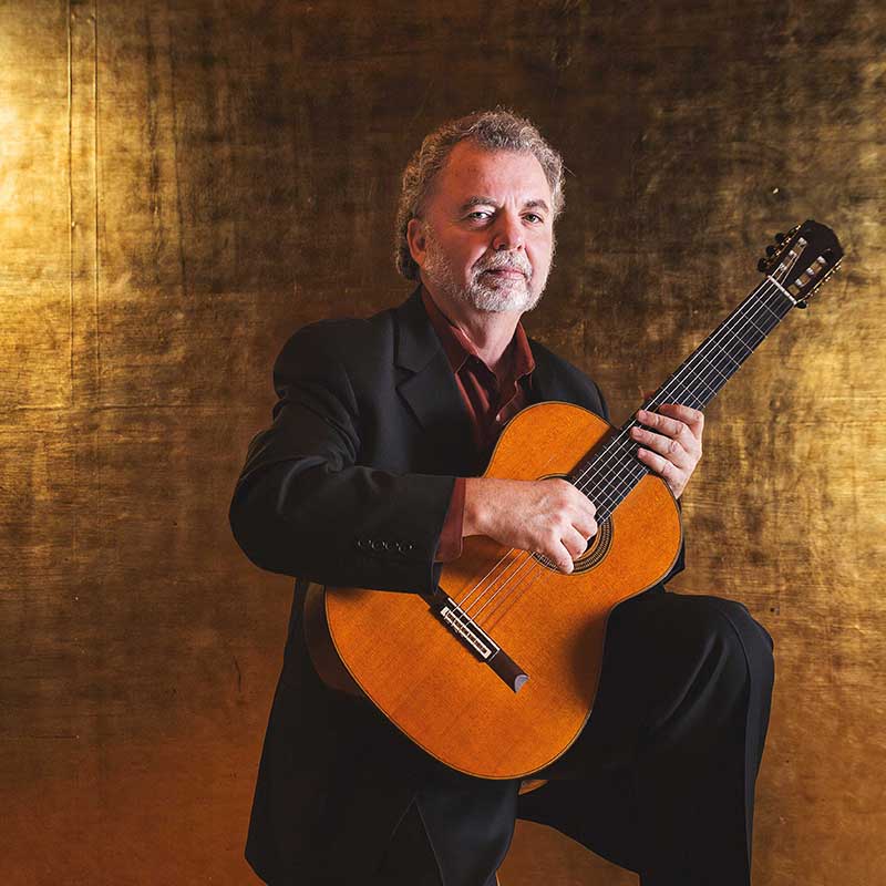 Manuel Barrueco standing with guitar in hand wearing black suit.