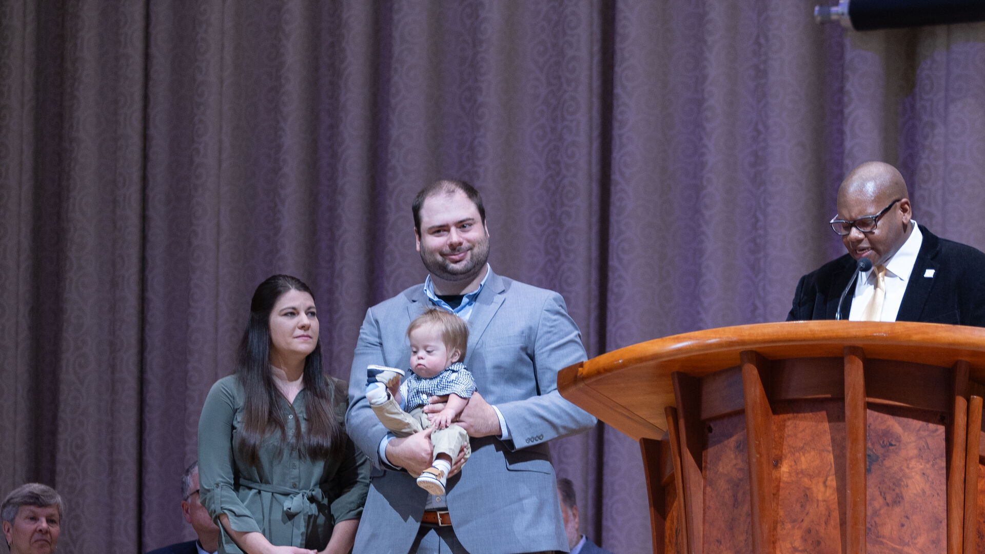 Houghton alumni Stephanie and David Bruno with their child standing as they are presented with an alumni award by President Lewis.