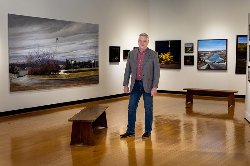 Houghton art professor John Rhett standing in the Ortlip Gallery in front of his paintings.