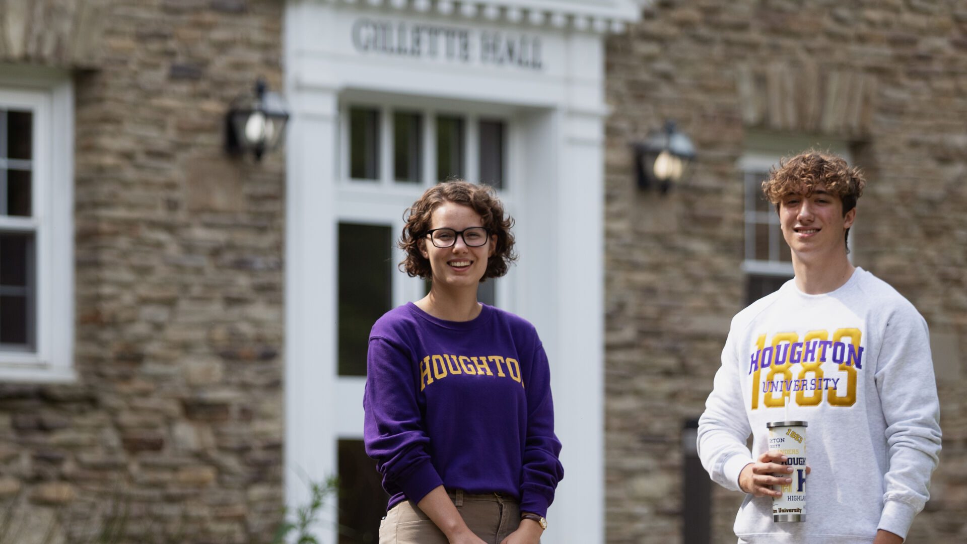 Two Houghton students standing in front of Gillette Hall wearing Houghton gear.