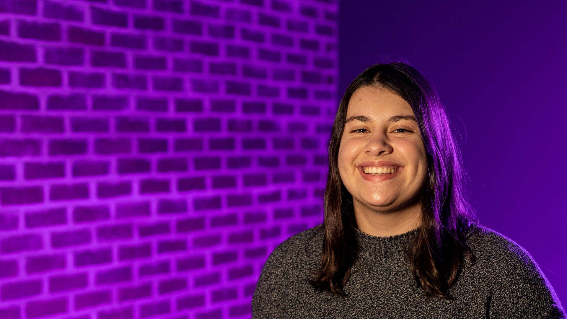 Houghton alumna Izzy Murch standing in front of purple tinted brick wall.