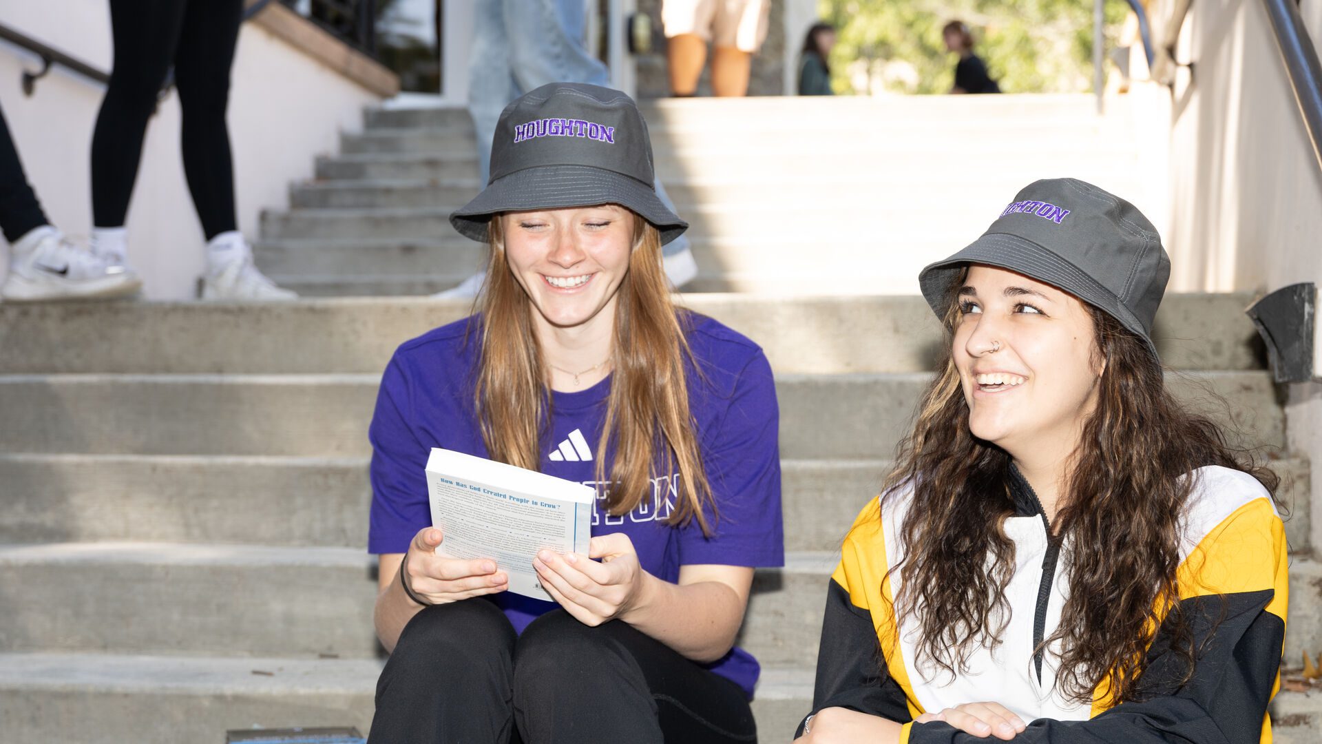 Two Houghton students sitting on the steps wearing Houghton gear and Houghton bucket hats.