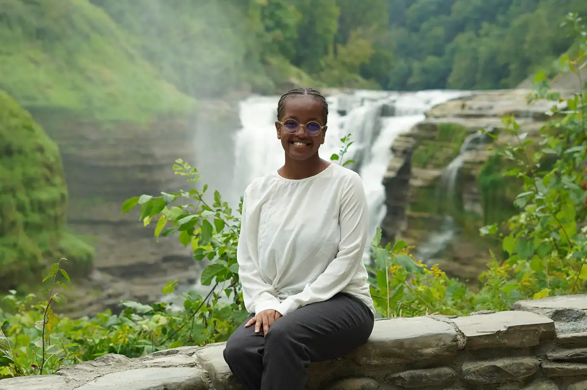 Houghton interconnect international student sitting on rock ledge in front of scenic waterfall.