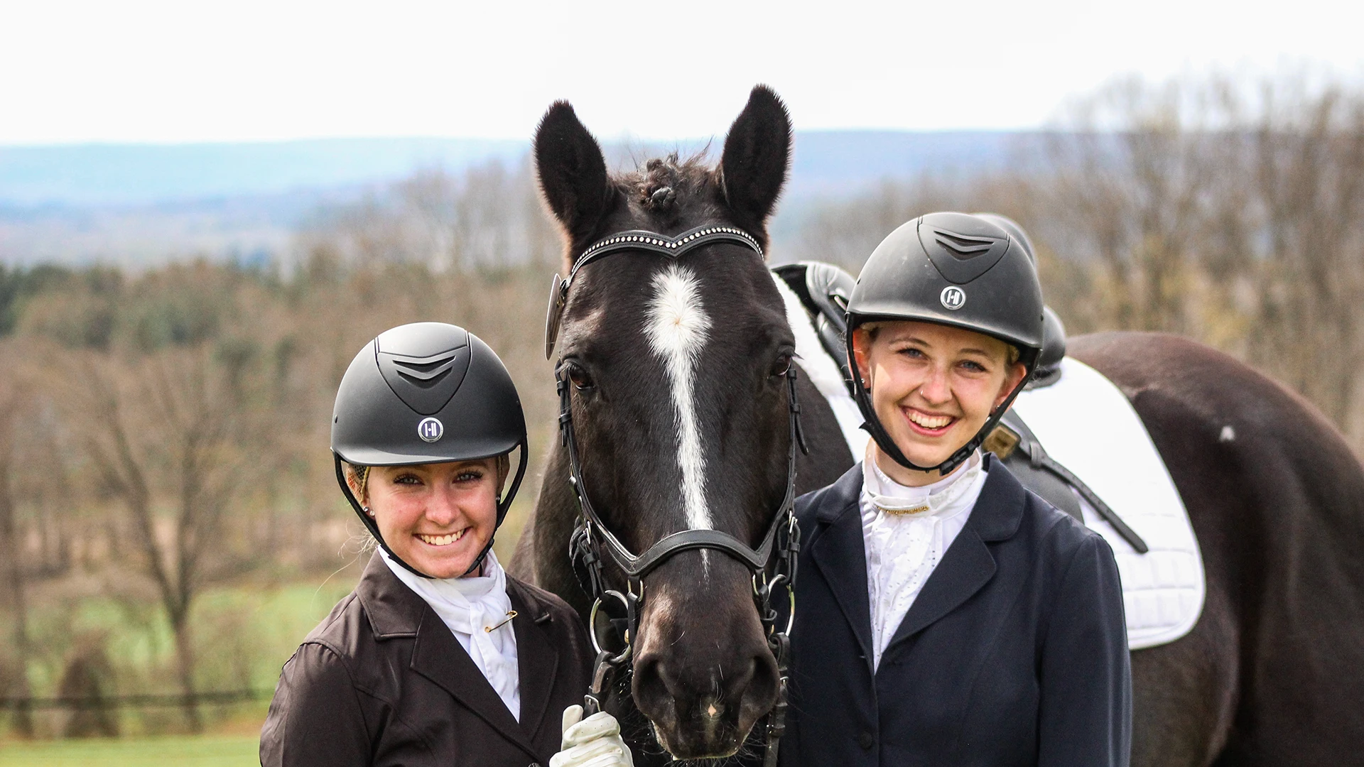Two Houghton equestrian students with helmets and show clothes standing next to black lesson horse outside.