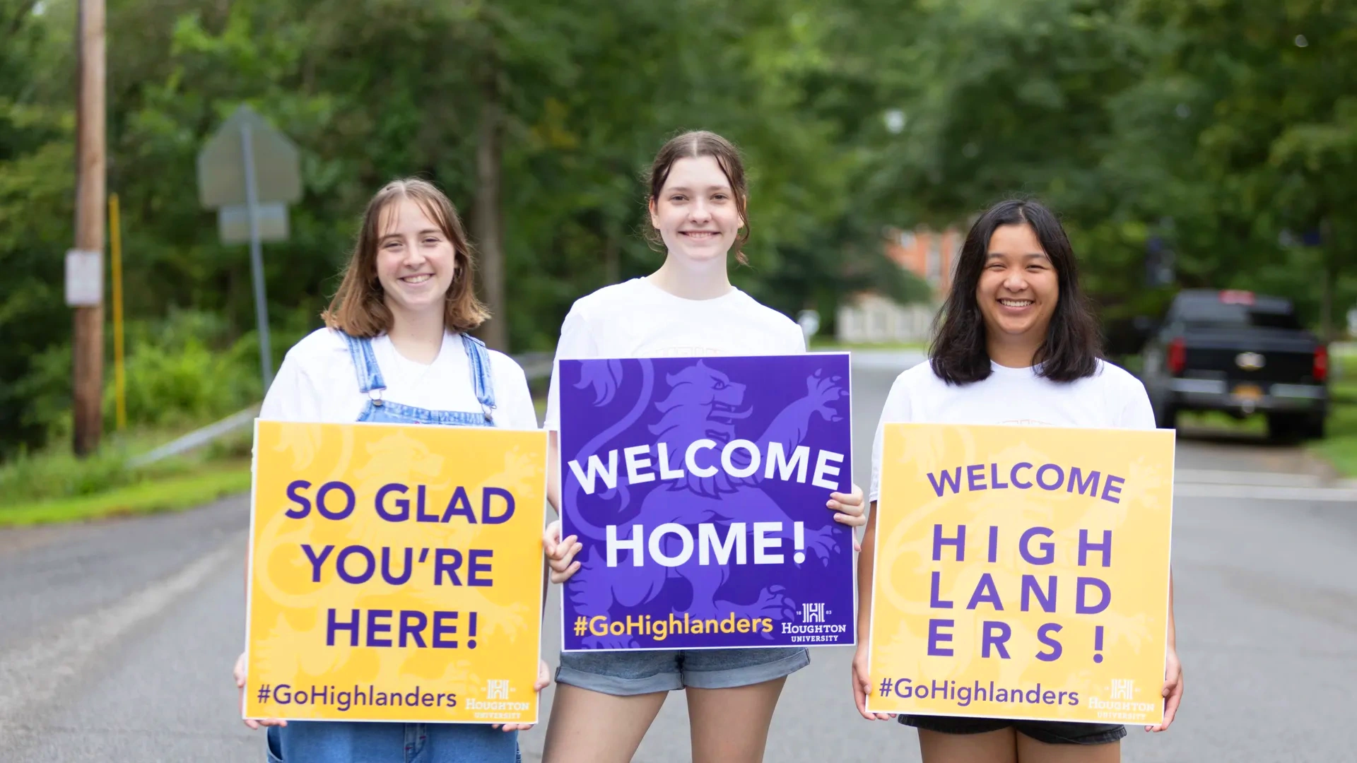 Three Houghton students welcoming new students with large gold and purple signs during move-in day.