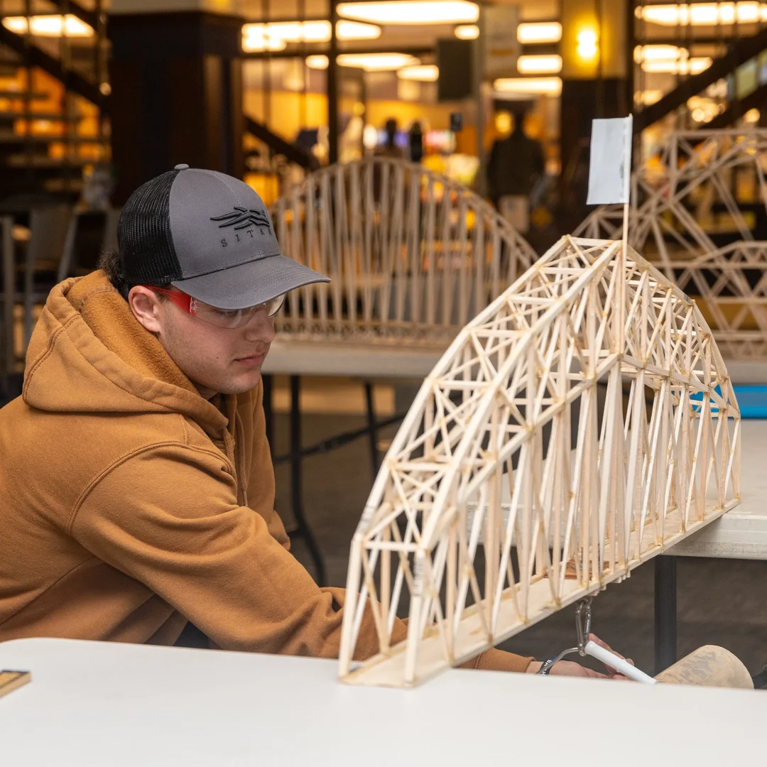 Houghton physics student testing bridge between two tables in campus center.