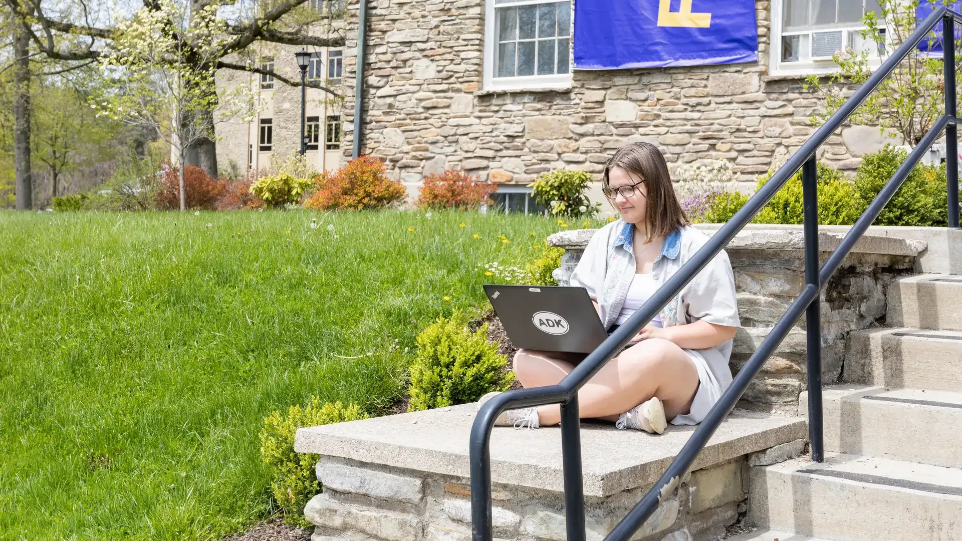 Houghton early college student sitting on steps working on coursework on laptop.