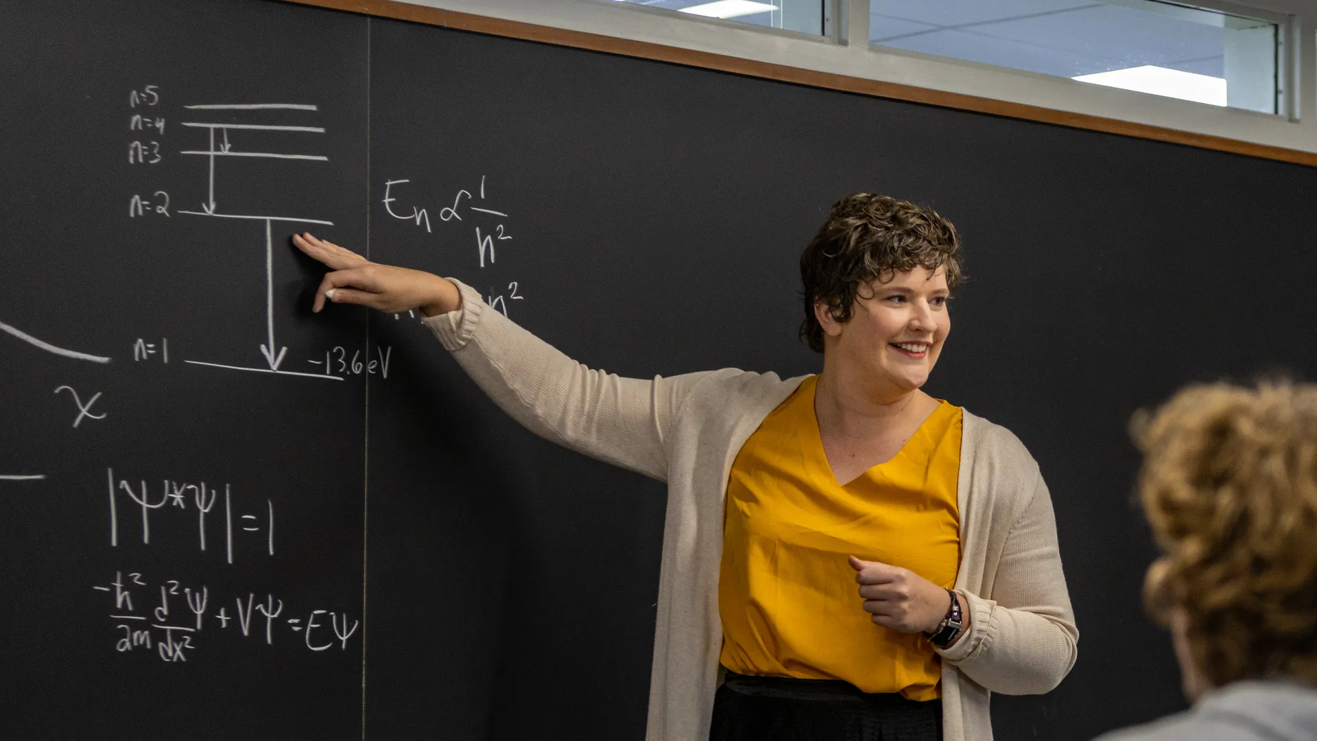 Houghton physics professor Katrina Koehler pointing at equation on blackboard.