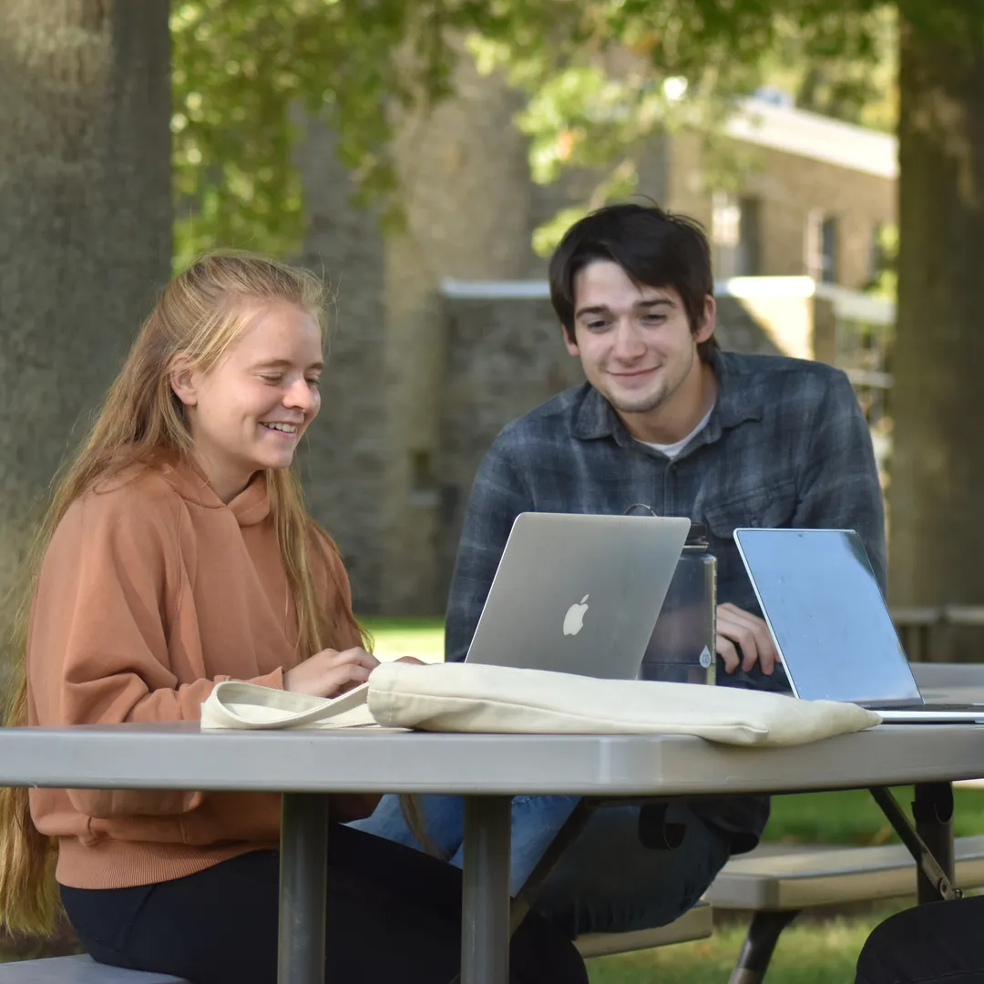 Two Houghton students sitting at outdoor reviewing work on their laptops together.