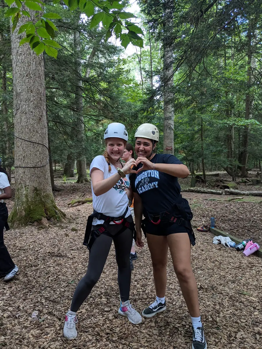 Two Houghton interconnect students wearing helmets on ropes course smiling and making heart with hands.