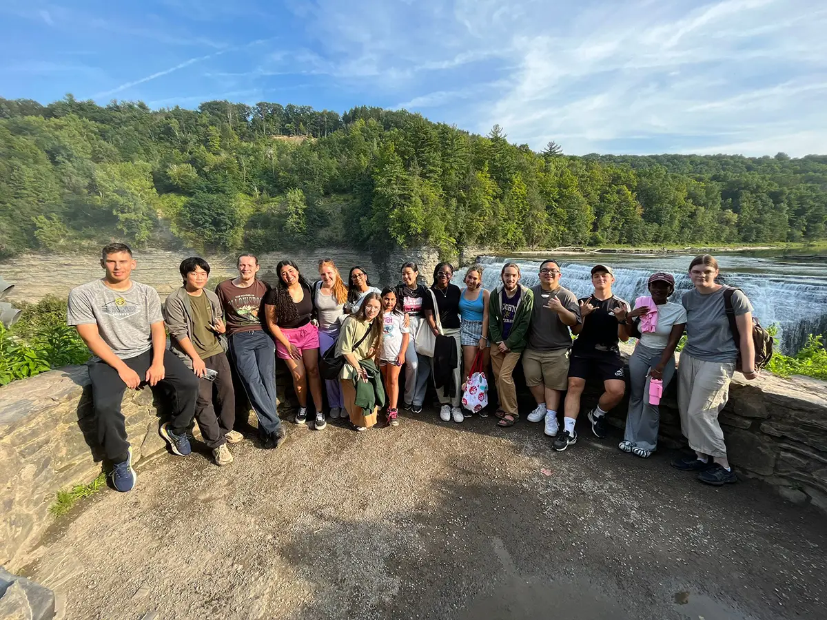 Houghton University interconnect students standing on by stonewall in front of waterfall.
