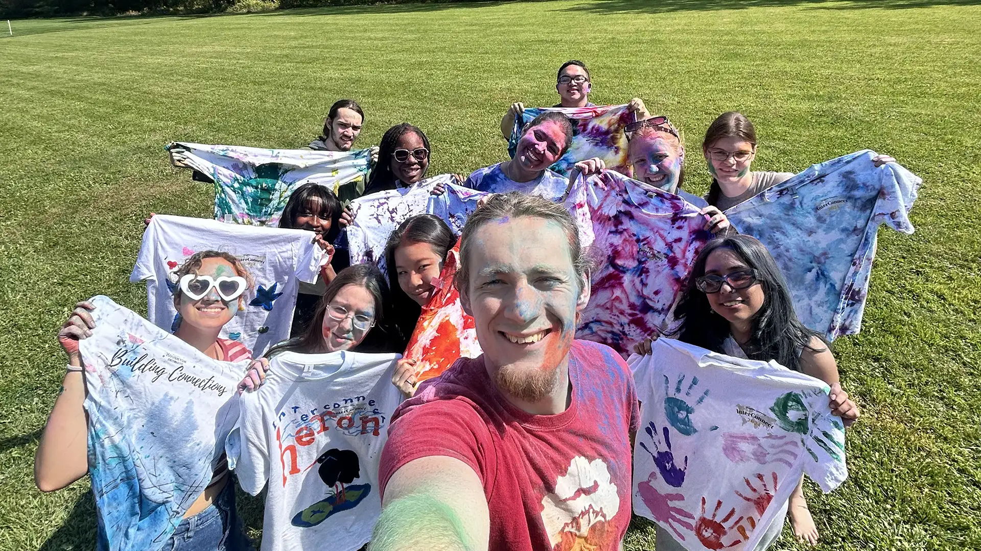 Houghton University interconnect students standing together covered in powdered color holding tie dye shirts.