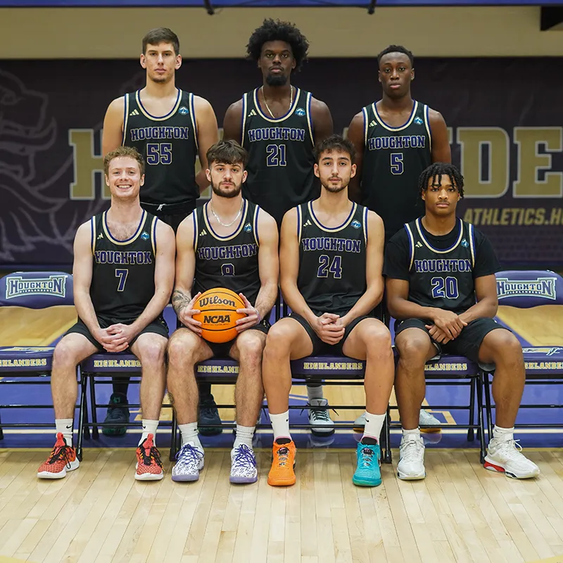 Houghton University Mens Basketball team sitting on the court smiling.