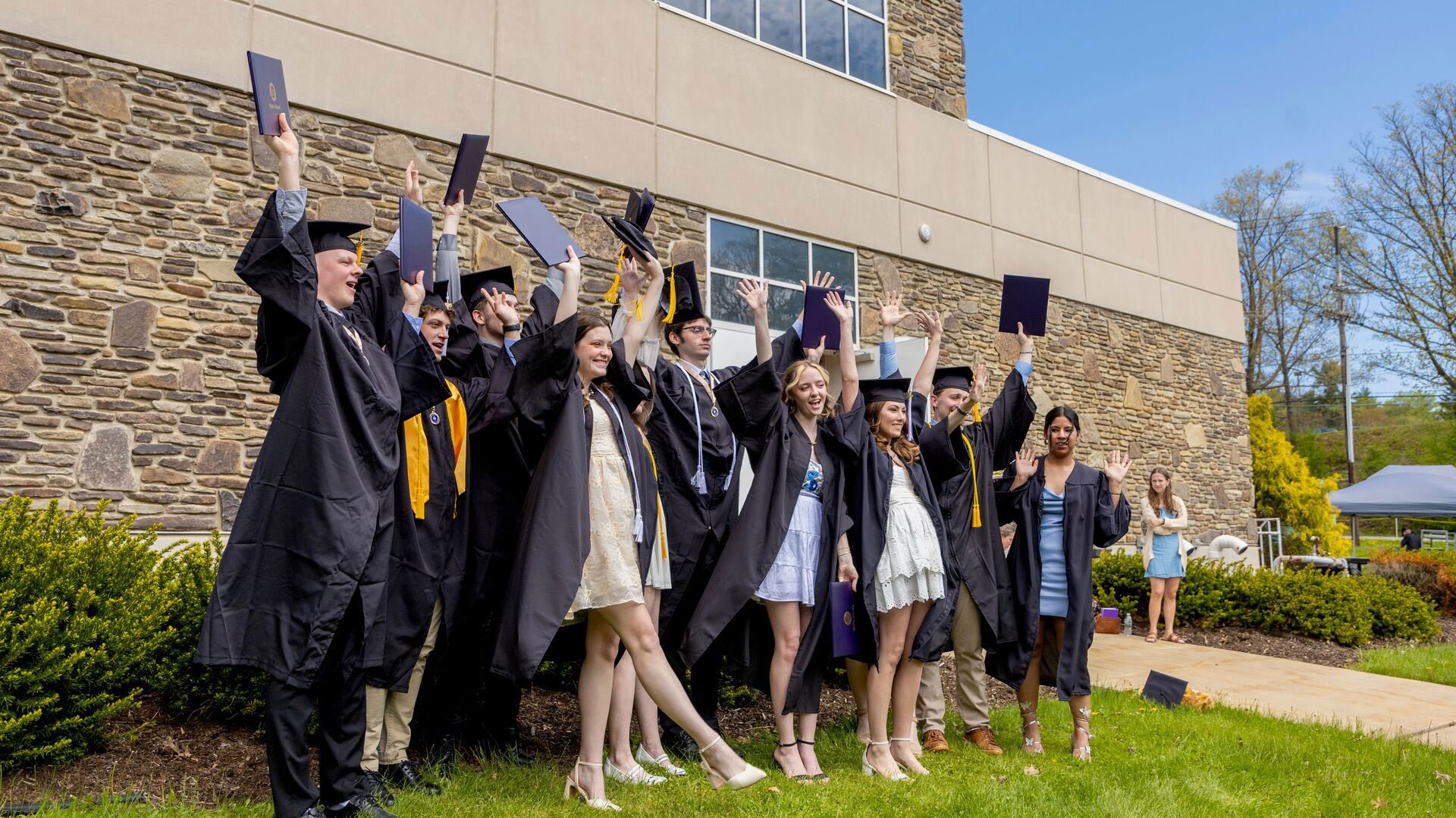 Group of graduates in caps and gowns celebrating outdoors, raising their arms in the air while holding diplomas. They're standing on a grassy area in front of a stone building with large windows. Students are wearing various colors underneath their black gowns and appear to be posing for a group photo.