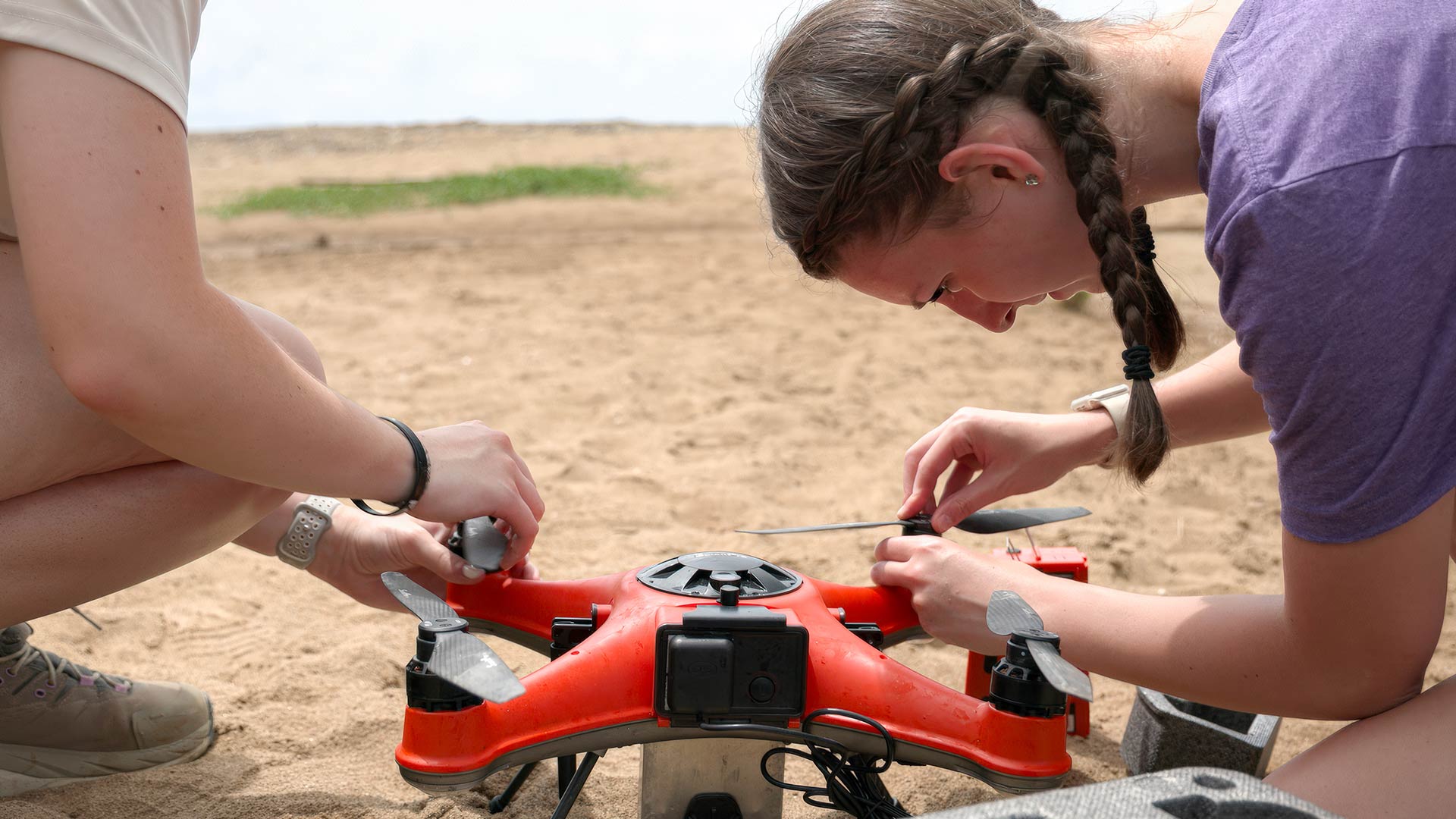 Two students prep an orange drone on a beach in Puerto Rico on a sunny day