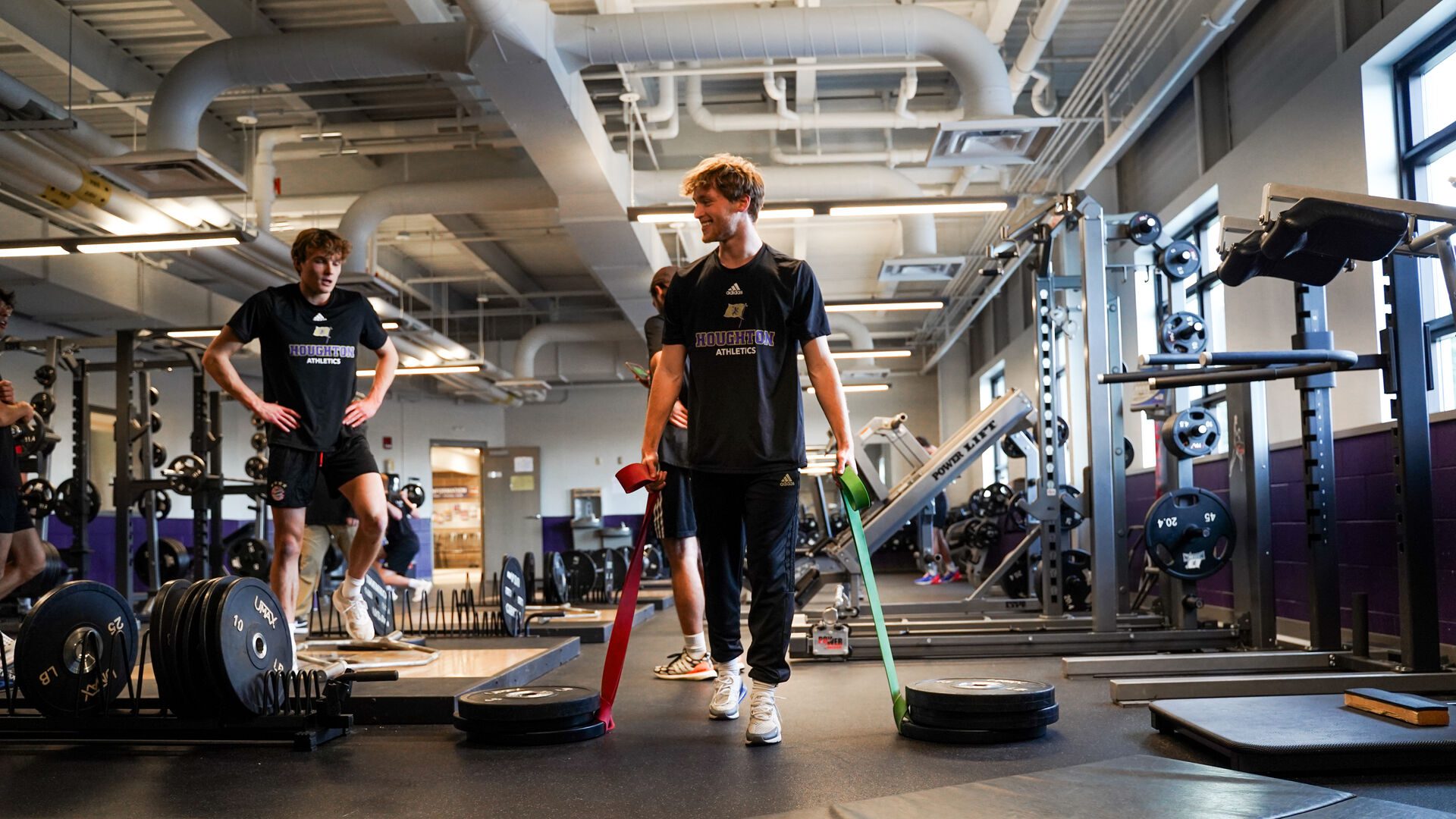 Houghton University student athletes in Houghton Athletics shirts working out in a modern gym facility. One student is performing resistance band exercises while another stands on a platform, surrounded by weight equipment and exercise machines